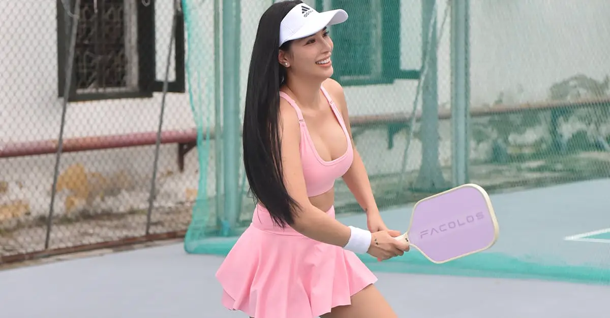A woman in a pink athletic outfit and white visor plays pickleball on an outdoor court, holding a paddle and smiling.