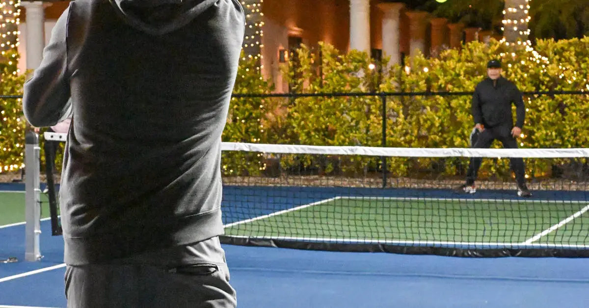 Two people are playing tennis on an outdoor court at night, with one person near the net and the other at the baseline. String lights and bushes are in the background.