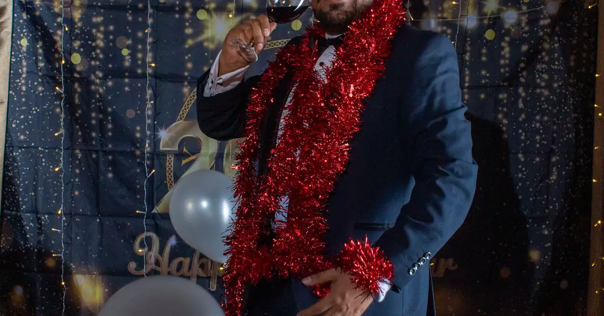 A man in a suit with a red tinsel garland holds a wine glass at a festive event, standing in front of a decorated backdrop with balloons.