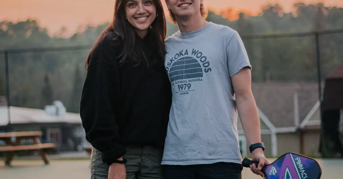 Two people standing on an outdoor court at sunset, one holding a pickleball paddle, both smiling at the camera. Trees and buildings are visible in the background.