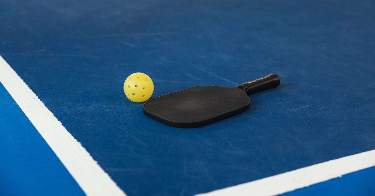 A black pickleball paddle and a yellow perforated pickleball resting on a blue court near white boundary lines.