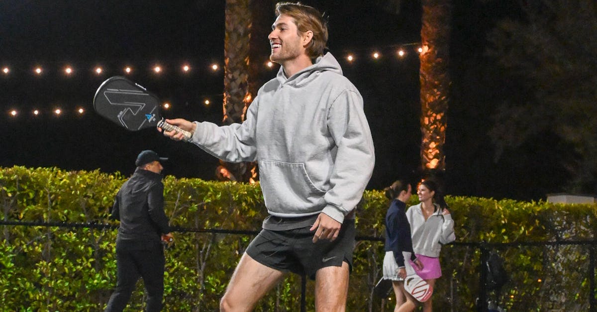 A man in a gray hoodie and black shorts plays pickleball outdoors at night; other people are visible in the background.
