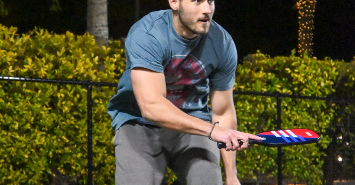 A man in a blue t-shirt and gray pants preparing to hit a ball with a paddle during a nighttime outdoor game.