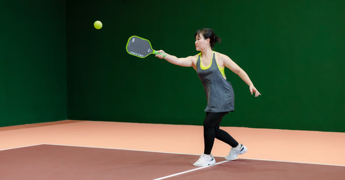 A woman in athletic wear hits a pickleball with a paddle on an indoor court with green walls and a tan floor.