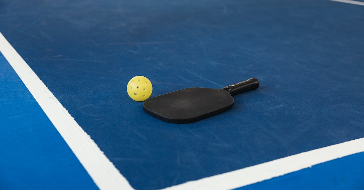 A black pickleball paddle and a yellow perforated ball rest on a blue court near a white boundary line.