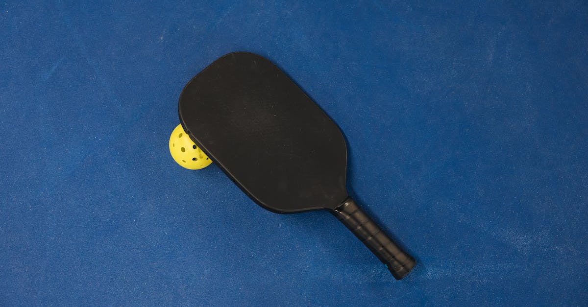 A black pickleball paddle rests on a blue court next to a yellow pickleball.
