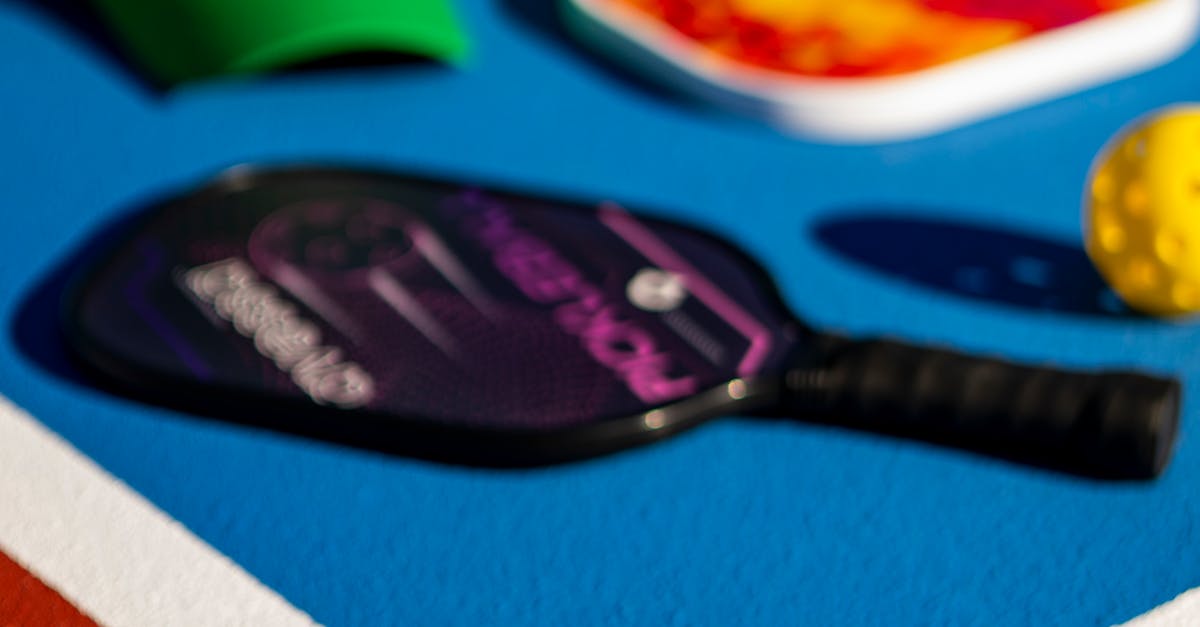 A close-up of a black and purple pickleball paddle lying on a blue court next to a yellow pickleball and other sports equipment.