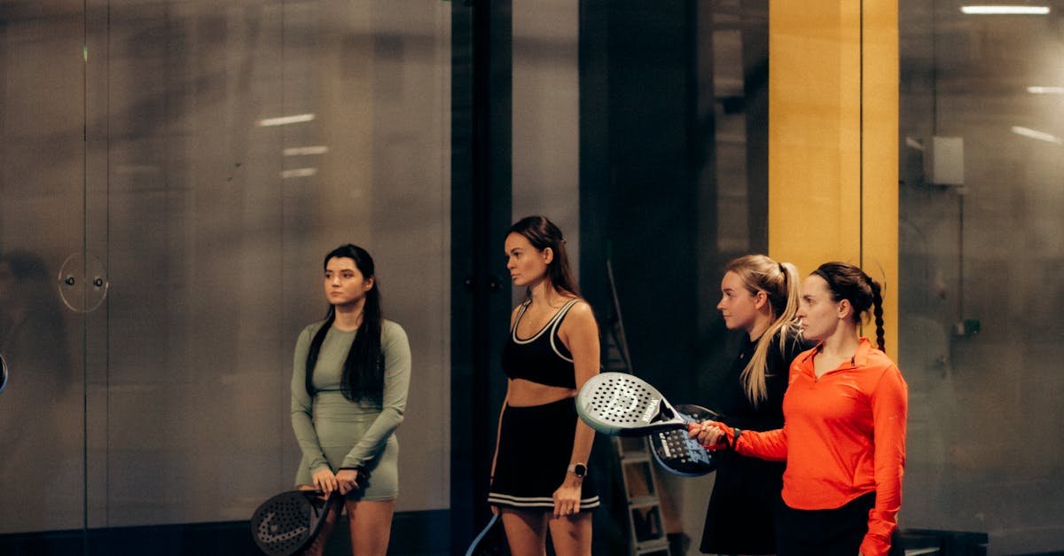 Four women standing indoors on a padel court, holding paddles and wearing sportswear, facing forward with neutral expressions.