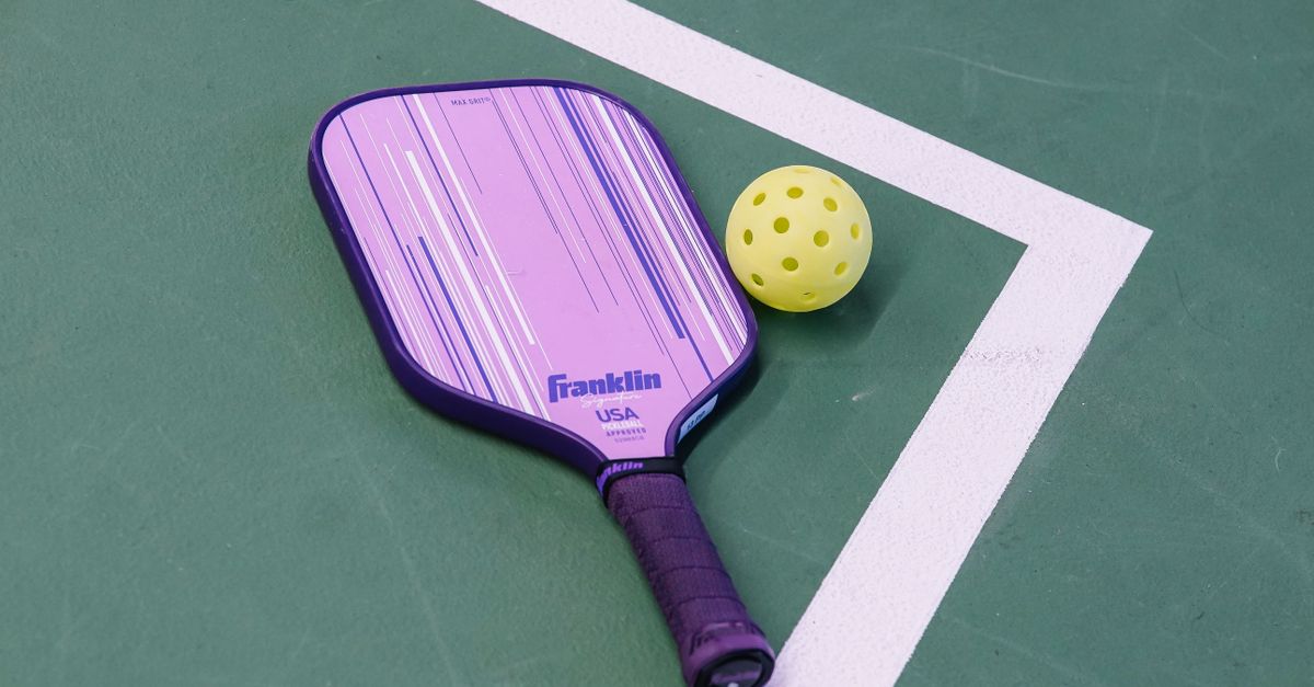 A purple Franklin pickleball paddle and a yellow pickleball resting on a green court near a white boundary line.