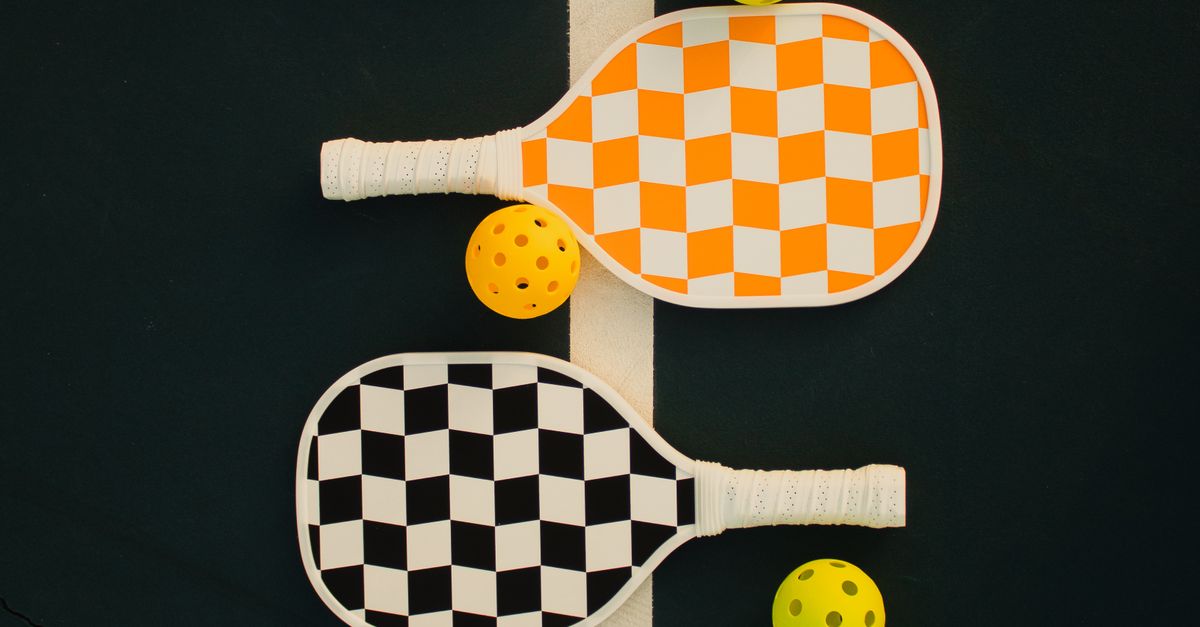 Two pickleball paddles with checkered designs, one orange and one black, and three yellow pickleballs are arranged on a dark court next to a white line.
